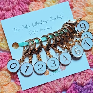 May include: Gold-coloured stitch markers with white enamel faces, numbered 0 to 9. The markers are attached to a light blue ribbon and a card that reads "The Cat's Whiskers Crochet Stitch Markers."