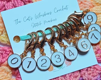 May include: A set of gold-colored stitch markers with white enamel faces, each numbered from 0 to 9. The markers are attached to a light blue ribbon and a card that reads "The Cat's Whiskers Crochet Stitch Markers."