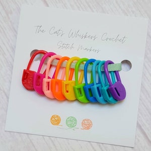 Puede incluir: Un conjunto de 10 marcadores de punto de plástico de colores en un patrón de arcoíris. Los marcadores tienen forma de bucle con cierre de botón. Los marcadores se muestran en una tarjeta blanca con el texto "The Cat's Whiskers Crochet Stitch Markers".