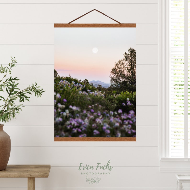 May include: A photograph of a sunset over a mountain range with a full moon in the sky. The foreground is a field of purple wildflowers.