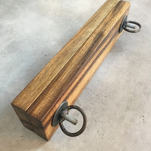 May include: A rectangular wooden object with a natural wood grain finish. It has metal ring attachments on each end, secured with bolts and washers. The wood has a warm, brown tone.