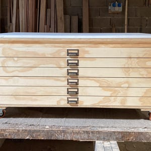 May include: A rectangular wooden storage cabinet featuring six horizontal drawers with light wood grain patterns. Each drawer is equipped with a dark metal label holder handle. The cabinet is supported by four small swivel castors at the base.
