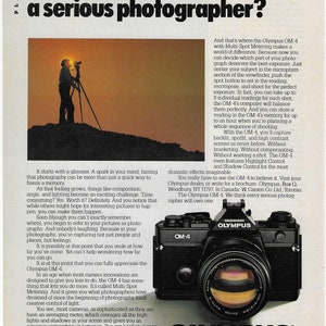 May include: A black Olympus OM-4 camera with a silver lens. The camera has a black strap and the text "Olympus" is printed in white on the camera body. The text "OM-4" is printed in white on the camera body. The text "WHEN YOU HAVE MORE TO SAY THAN JUST SMILE" is printed in white below the camera. The text "OLYMPUS" is printed in white above the camera.