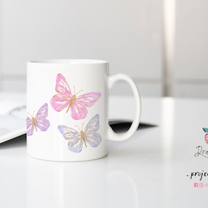 Puede incluir: Taza de cer&aacute;mica blanca con tres mariposas pintadas en ella. Las mariposas son de color rosa, morado y azul con detalles de purpurina dorada.