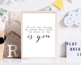 May include: A framed print with a white background and black text that reads "Of all the things my hands have held, the best so far is you". The print is on a white shelf with other decorative items, including a stuffed animal, a wooden house, and a light-up cloud.