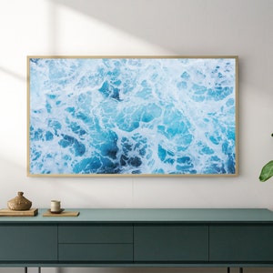 K&ouml;nnte beinhalten: Ein gerahmter Wandkunstdruck mit einer Luftaufnahme von Meereswellen in Blau- und Wei&szlig;t&ouml;nen. Das Kunstwerk wird &uuml;ber einem t&uuml;rkisfarbenen Schrank mit einer dekorativen Vase und einer Pflanze im Vordergrund pr&auml;sentiert.