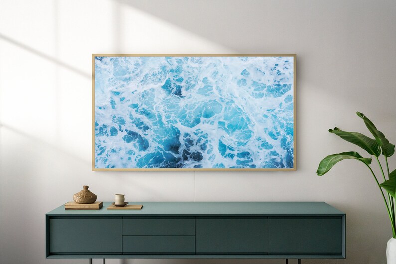 K&ouml;nnte beinhalten: Ein gerahmter Wandkunstdruck mit einer Luftaufnahme von Meereswellen in Blau- und Wei&szlig;t&ouml;nen. Das Kunstwerk wird &uuml;ber einem t&uuml;rkisfarbenen Schrank mit einer dekorativen Vase und einer Pflanze im Vordergrund pr&auml;sentiert.