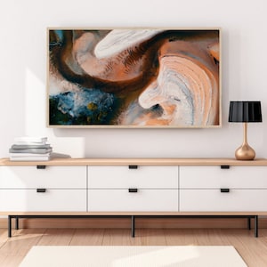 May include: A framed abstract art print with swirling patterns in shades of brown, orange, white, and blue. The print is hanging on a white wall above a wooden dresser with white drawers and black handles.