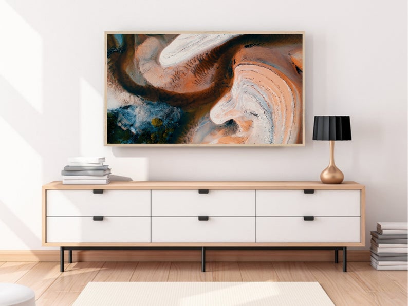 May include: A framed abstract art print with swirling patterns in shades of brown, orange, white, and blue. The print is hanging on a white wall above a wooden dresser with white drawers and black handles.