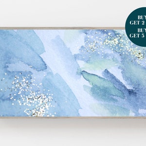 Puede incluir: Pintura abstracta de acuarela con tonos azules y verdes y purpurina plateada. La pintura está enmarcada en un marco de madera claro.