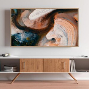 May include: A framed abstract art print with swirling patterns in shades of brown, white, blue, and green. The print is hanging on a white wall above a white and wood cabinet.
