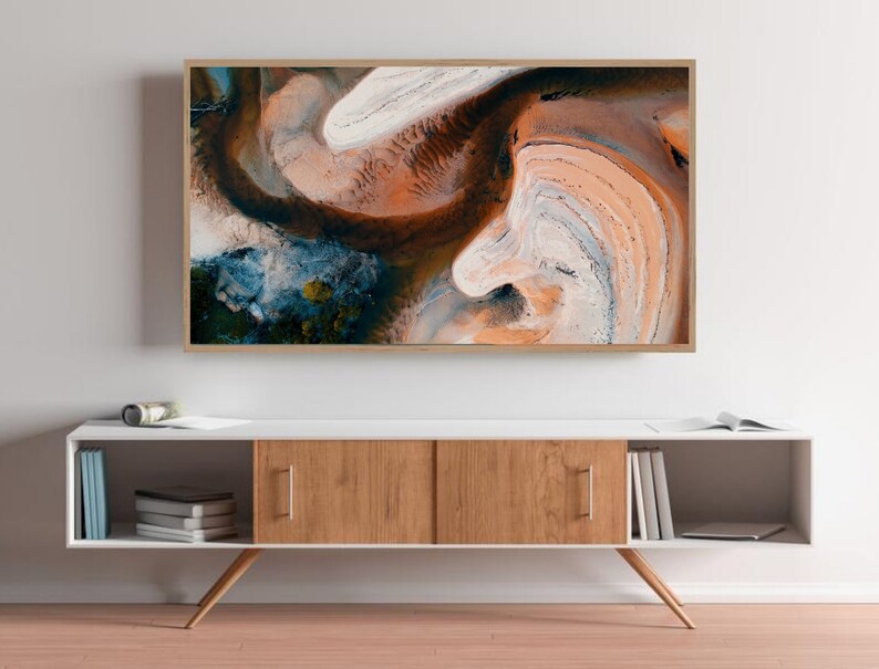 May include: A framed abstract art print with swirling patterns in shades of brown, white, blue, and green. The print is hanging on a white wall above a white and wood cabinet.