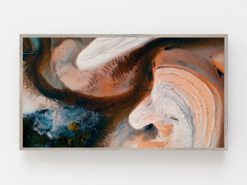 May include: Abstract aerial photograph of a landscape with swirling patterns of orange, brown, white, and blue. The image is framed in a light wood frame.