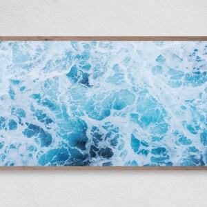 K&ouml;nnte beinhalten: Gerahmter Druck mit einer Luftaufnahme von Meereswellen. Das Bild zeigt ein dynamisches Muster aus t&uuml;rkisem und wei&szlig;em Wasser, das einen strukturierten Effekt erzeugt. Der Druck ist in einem hellbraunen Holzrahmen eingefasst, geeignet f&uuml;r Wanddekoration.