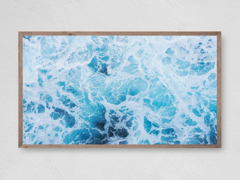 K&ouml;nnte beinhalten: Gerahmter Druck mit einer Luftaufnahme von Meereswellen. Das Bild zeigt ein dynamisches Muster aus t&uuml;rkisem und wei&szlig;em Wasser, das einen strukturierten Effekt erzeugt. Der Druck ist in einem hellbraunen Holzrahmen eingefasst, geeignet f&uuml;r Wanddekoration.