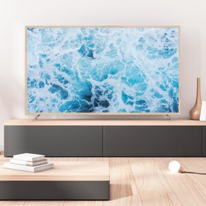 K&ouml;nnte beinhalten: Ein modernes Wohnzimmer-Setup mit einem gro&szlig;en Flachbildfernseher, der ein Bild von Meereswellen zeigt. Der Fernseher hat einen hellen Holzrahmen. Eine Holzgitarre steht links. Ein niedriger, zweifarbiger Schrank enth&auml;lt dekorative Gegenst&auml;nde. Ein kleiner Couchtisch und eine Gl&uuml;hbirne sind ebenfalls vorhanden.