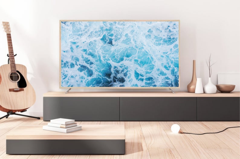 K&ouml;nnte beinhalten: Ein modernes Wohnzimmer-Setup mit einem gro&szlig;en Flachbildfernseher, der ein Bild von Meereswellen zeigt. Der Fernseher hat einen hellen Holzrahmen. Eine Holzgitarre steht links. Ein niedriger, zweifarbiger Schrank enth&auml;lt dekorative Gegenst&auml;nde. Ein kleiner Couchtisch und eine Gl&uuml;hbirne sind ebenfalls vorhanden.