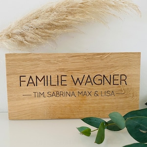 Könnte beinhalten: Ein Holzschild mit dem Text "FAMILIE WAGNER - TIM, SABRINA, MAX & LISA -" in dunkelbrauner Farbe graviert.