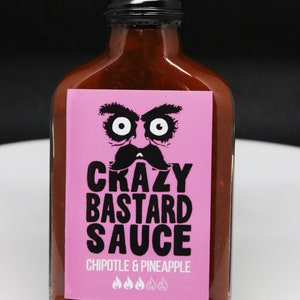 May include: A glass bottle of "Crazy Bastard Sauce" with a pink label featuring a cartoon face with a mustache. The label reads "Chipotle & Pineapple" and has four chili pepper icons.