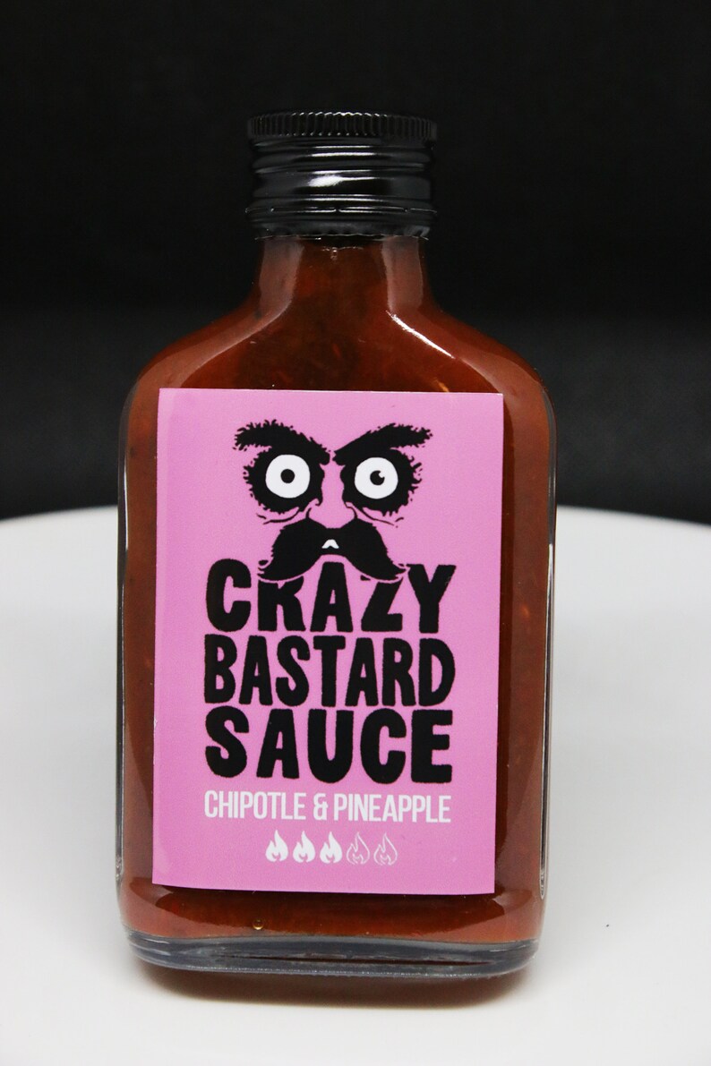 May include: A glass bottle of "Crazy Bastard Sauce" with a pink label featuring a cartoon face with a mustache. The label reads "Chipotle & Pineapple" and has four chili pepper icons.