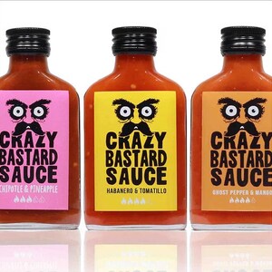 May include: Three glass bottles of hot sauce with black lids and labels. The labels have a cartoon face with a mustache and the text "Crazy Bastard Sauce". The flavors are Chipotle & Pineapple, Habanero & Tomatillo, and Ghost Pepper & Mango.
