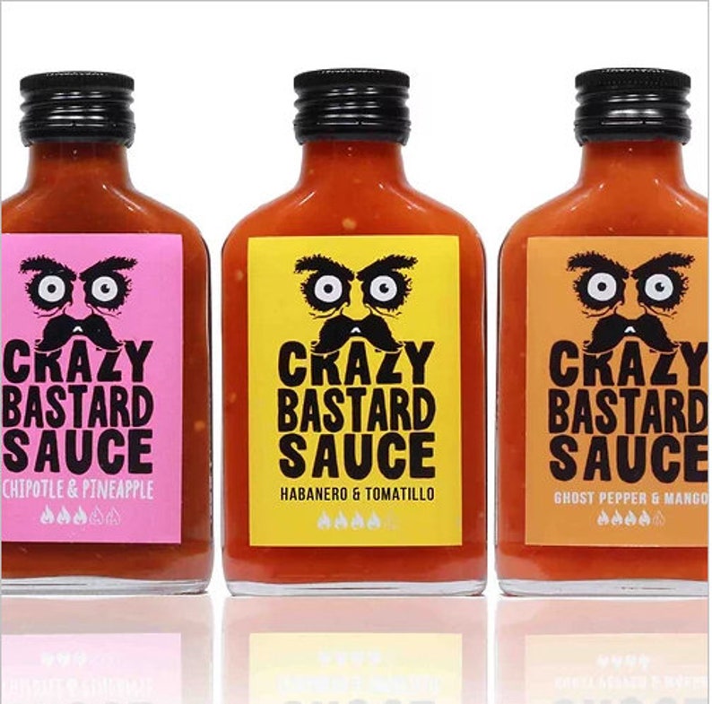 May include: Three glass bottles of hot sauce with black lids and labels. The labels have a cartoon face with a mustache and the text "Crazy Bastard Sauce". The flavors are Chipotle & Pineapple, Habanero & Tomatillo, and Ghost Pepper & Mango.