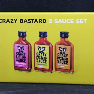 May include: A yellow box with the text "CRAZY BASTARD 3 SAUCE SET" on the front. Three glass bottles of hot sauce are displayed on the front of the box. The bottles are labeled "CRAZY BASTARD SAUCE" with different flavors: "CHILLI & PINEAPPLE", "HABANERO & TOMATILLO", and "CHILLI PEPPER & GARLIC".