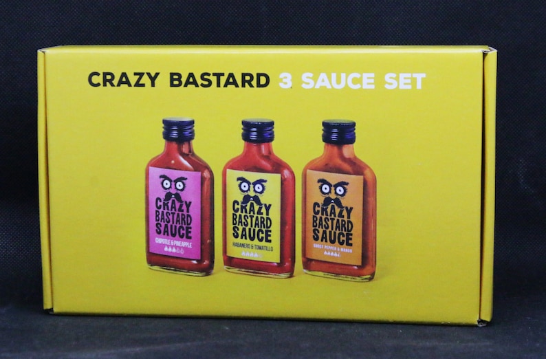 May include: A yellow box with the text "CRAZY BASTARD 3 SAUCE SET" on the front. Three glass bottles of hot sauce are displayed on the front of the box. The bottles are labeled "CRAZY BASTARD SAUCE" with different flavors: "CHILLI & PINEAPPLE", "HABANERO & TOMATILLO", and "CHILLI PEPPER & GARLIC".
