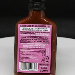 May include: A 100ml bottle of smoked chili sauce with a pink label. The label reads "Scharfe Sauce mit ger&auml;uchertem Chili" and includes the manufacturer's name, address, and nutritional information. The bottle has a black cap and is filled with a dark red sauce.