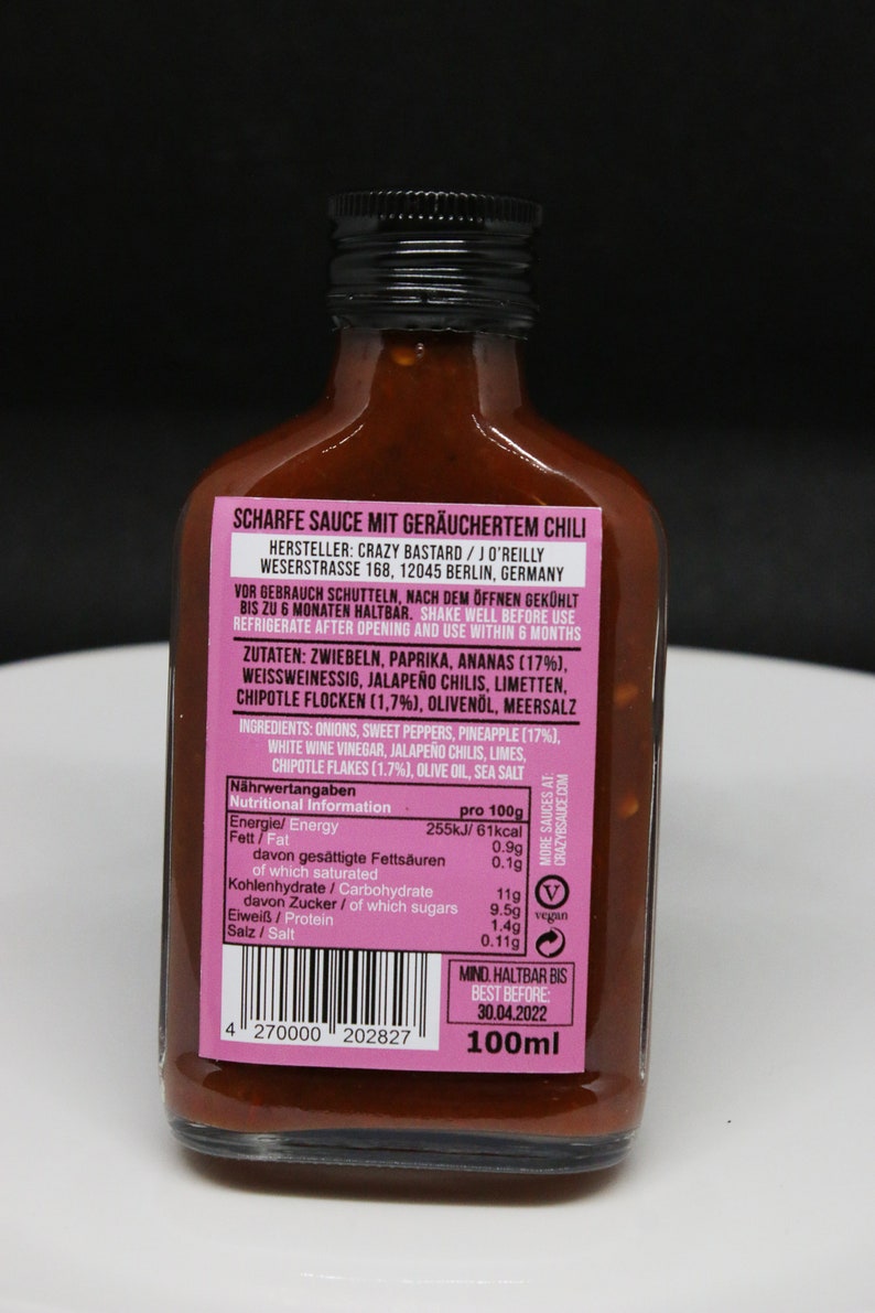 May include: A 100ml bottle of smoked chili sauce with a pink label. The label reads "Scharfe Sauce mit ger&auml;uchertem Chili" and includes the manufacturer's name, address, and nutritional information. The bottle has a black cap and is filled with a dark red sauce.