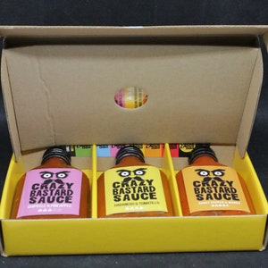 May include: A yellow cardboard box with three glass bottles of hot sauce inside. The bottles are labeled "Crazy Bastard Sauce" with different flavor combinations: Chipotle & Pineapple, Habanero & Tomatillo, and Ghost Pepper & Mango.