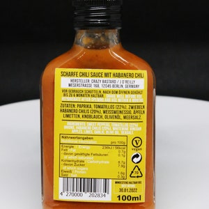May include: A 100ml bottle of hot chili sauce with a yellow label. The label reads "Scharfe Chili Sauce mit Habanero Chili" and lists the ingredients, nutritional information, and best by date. The bottle is on a black background.