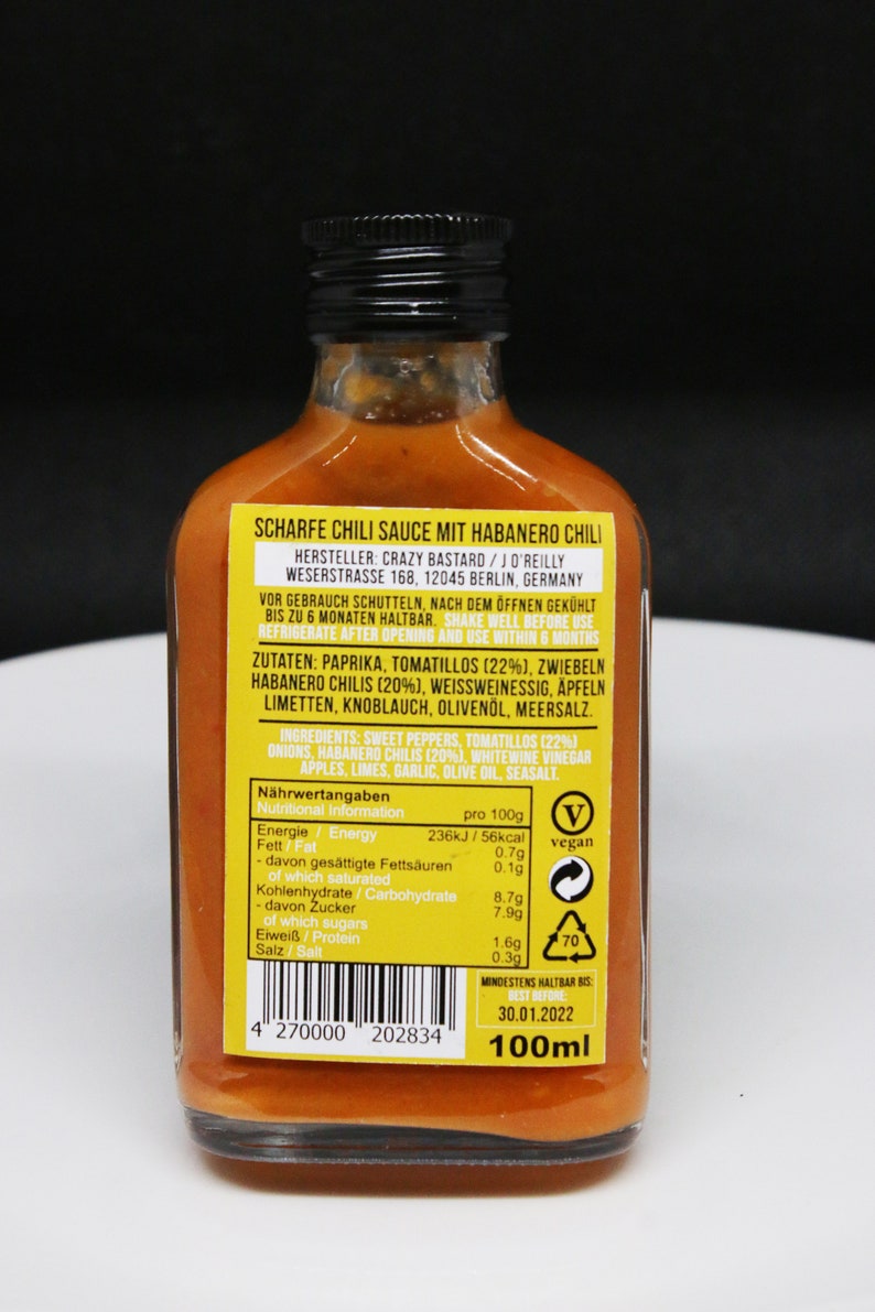 May include: A 100ml bottle of hot chili sauce with a yellow label. The label reads "Scharfe Chili Sauce mit Habanero Chili" and lists the ingredients, nutritional information, and best by date. The bottle is on a black background.