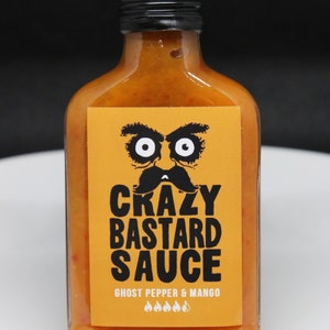May include: A glass bottle of orange hot sauce with a black label. The label has a cartoon face with a mustache and the text "Crazy Bastard Sauce" and "Ghost Pepper & Mango".