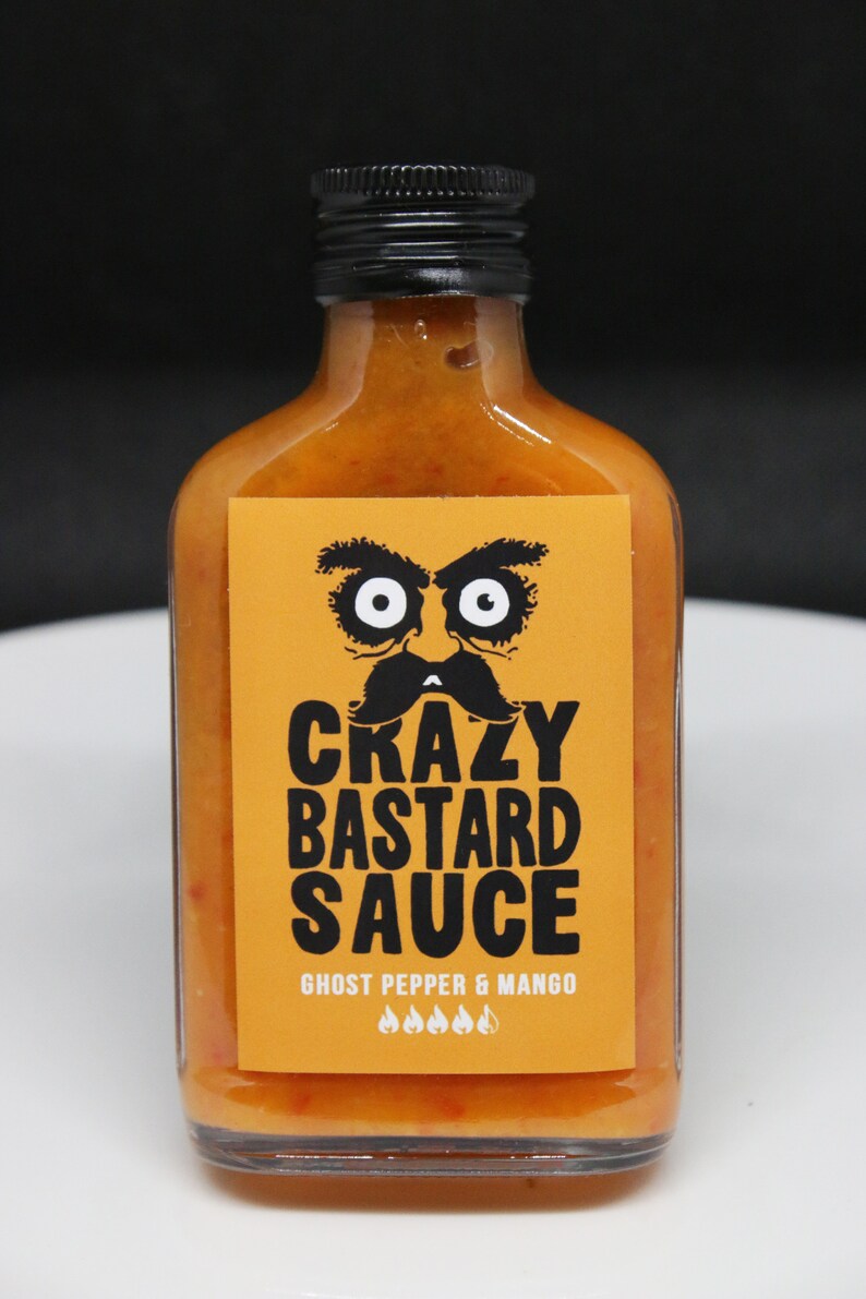 May include: A glass bottle of orange hot sauce with a black label. The label has a cartoon face with a mustache and the text "Crazy Bastard Sauce" and "Ghost Pepper & Mango".