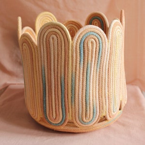 Può includere: Un cesto intrecciato con un bordo a festoni, realizzato con corda spessa in tonalità beige, giallo e blu.