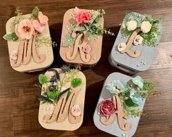 Puede incluir: Cinco pequeñas cajas de regalo con adornos florales y letras de purpurina. Las cajas están decoradas con flores de diferentes colores y están etiquetadas con las letras M, K, L, M y A.