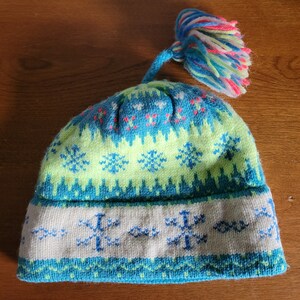 Peut inclure: Un bonnet d'hiver en tricot bleu, vert et blanc avec un pompon coloré sur le dessus. Le bonnet a un motif de flocons de neige.