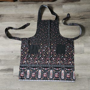 May include: Black apron with a floral pattern featuring pink, yellow, and white flowers. The apron has two pockets and a decorative border with framed floral designs. It includes a neck strap and waist ties.