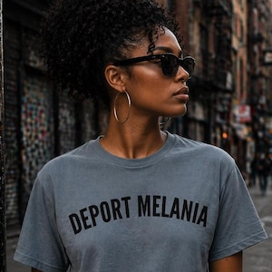 May include: A slate blue short-sleeved crew neck t-shirt featuring the text DEPORT MELANIA printed in black capital letters across the chest. The shirt is made of a solid-colored fabric and has a standard fit with a straight hemline.