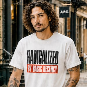 May include: A white short-sleeved t-shirt featuring the text RADICALIZED in bold black letters above the text BY BASIC DECENCY in white letters on a red rectangular background. The shirt is paired with dark charcoal grey denim jeans and black trainers.
