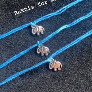 Puede incluir: Tres pulseras de cuerda azul con dijes de elefante plateados. Las pulseras están dispuestas en una fila sobre un fondo negro. El texto "Rakhis for kids" es visible en la parte superior de la imagen.