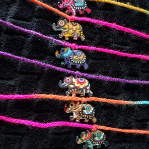 May include: A selection of decorative elephant charms in various colours, strung on brightly coloured yarn. The elephant charms feature detailed patterns and are set against a black, textured background. The yarn is in shades of orange, purple, and pink.