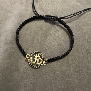 May include: A black string bracelet with a gold Om symbol charm. The charm is surrounded by small black stones.