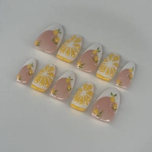 May include: A set of ten artificial nails with a lemon theme. The nails feature a white tip, a pink base, and yellow lemon slice and lemon sprig designs. The nails are oval-shaped and arranged on a white surface.