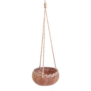 May include: A hanging planter made from a natural coconut shell with a rope hanger. The planter has a brown color and a woven design around the top edge.