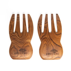 May include: Two wooden salad servers with a natural wood grain pattern. Each server has a wide, curved handle and three tines. The text "Rainforest Bowls" is printed on the handle of each server.