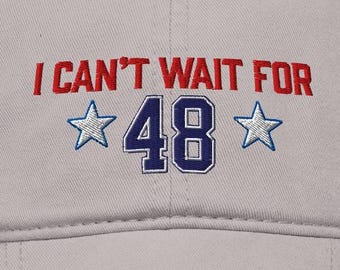 May include: A light gray baseball cap with the text "I CAN'T WAIT FOR" in red, and "48" in blue with two blue stars. The cap has a curved brim and a button on top.