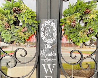 May include: A black wooden welcome sign with white lettering that reads "Welcome" and "The Knuckles Home". The sign is decorated with a wreath and pine cones.