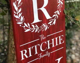 May include: Red wooden sign with a white wreath and the letter "R" in the center. The text "The Ritchie Family" is written below the wreath.